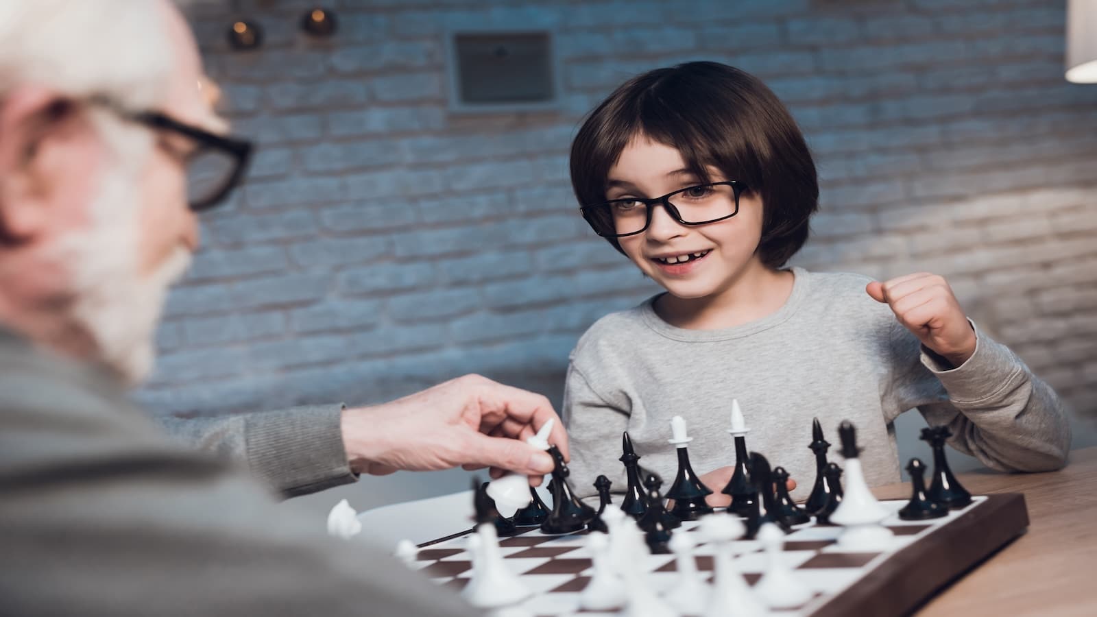 Grandfather and grandson are playing chess together at table at night at home.