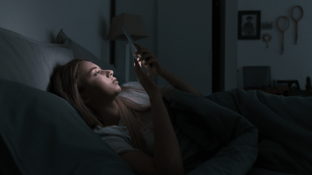 Young sleepy exhausted woman lying in bed using smartphone at late night with bluelight.