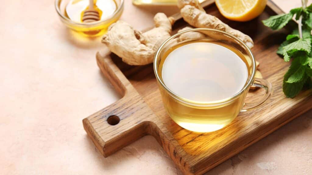 Glass cup of ginger tea and bowl with honey on pink background