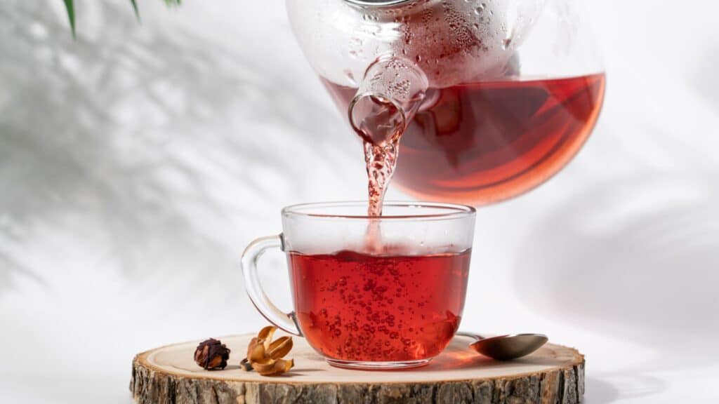 Hot tea is pouring from glass teapot into cup. Hibiscus red tea in glass cup close-up view.