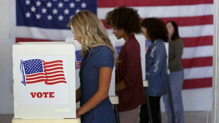 Four women of various demographics, young blonde woman in front, filling in ballots and casting votes in booths at polling station, US flag on wall at back. Focus on booth signage