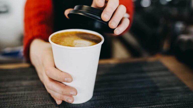 Barista in apron is holding in hands hot cappuccino in white takeaway paper cup. Coffee take away at cafe shop