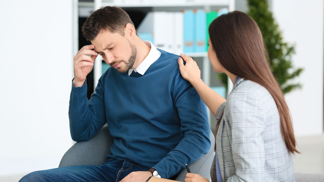 Man sitting in a seat, being comforted by an outstretched hand from a Female psychologist with patient in office.