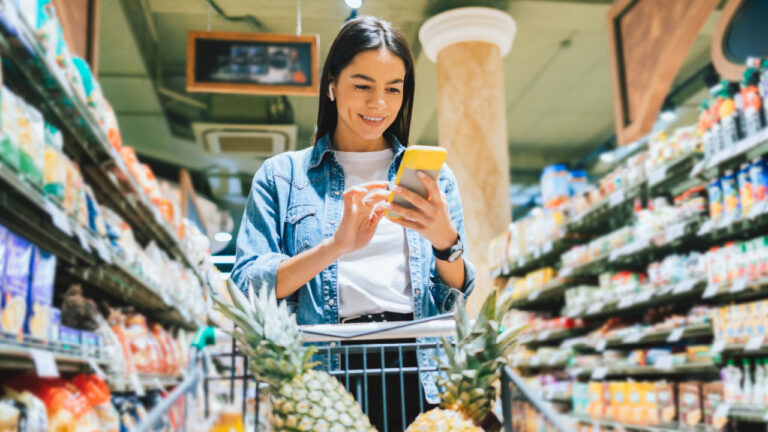 Happy young woman wearing earphones using smartphone checklist in supermarket