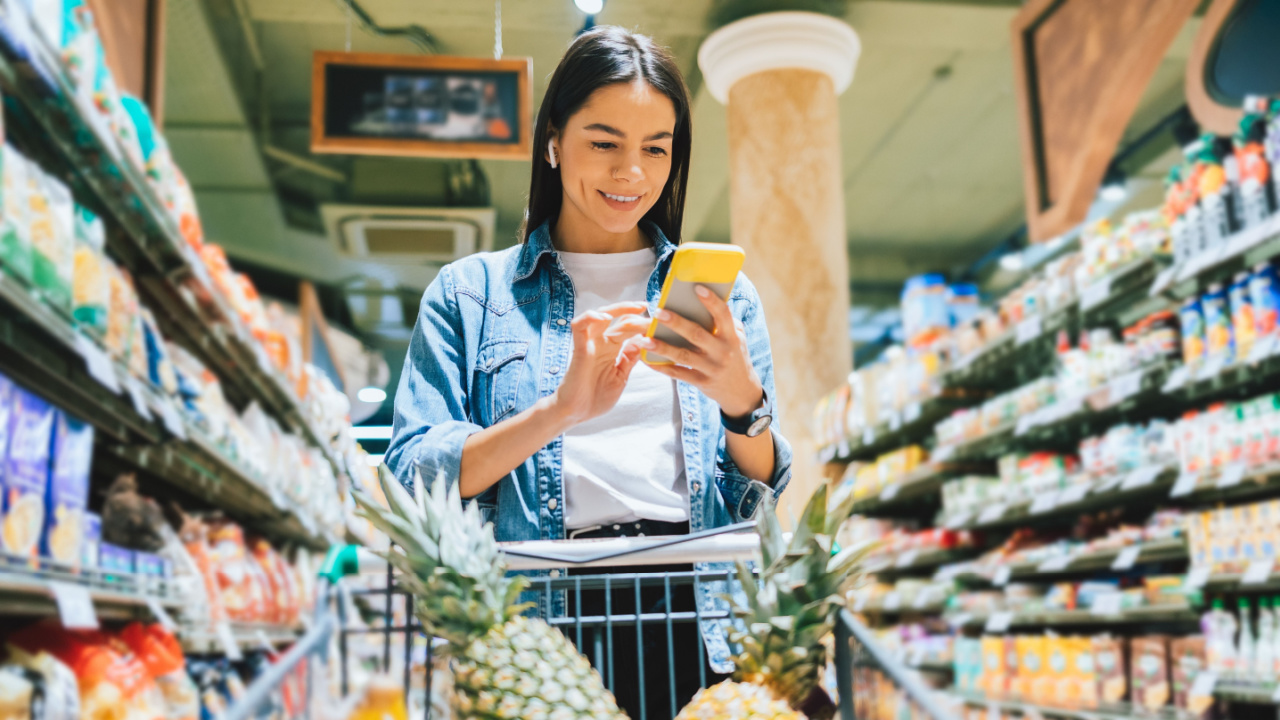Happy young woman wearing earphones using smartphone checklist in supermarket