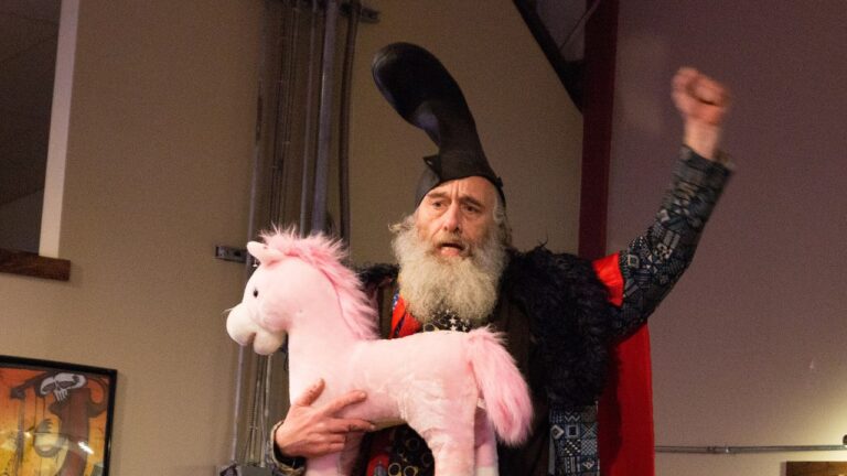 Vermin Supreme raising his hand in victory after a public appearance. He is wearing a large boot on his head and holding a stuffed pink unicorn with his other arm.