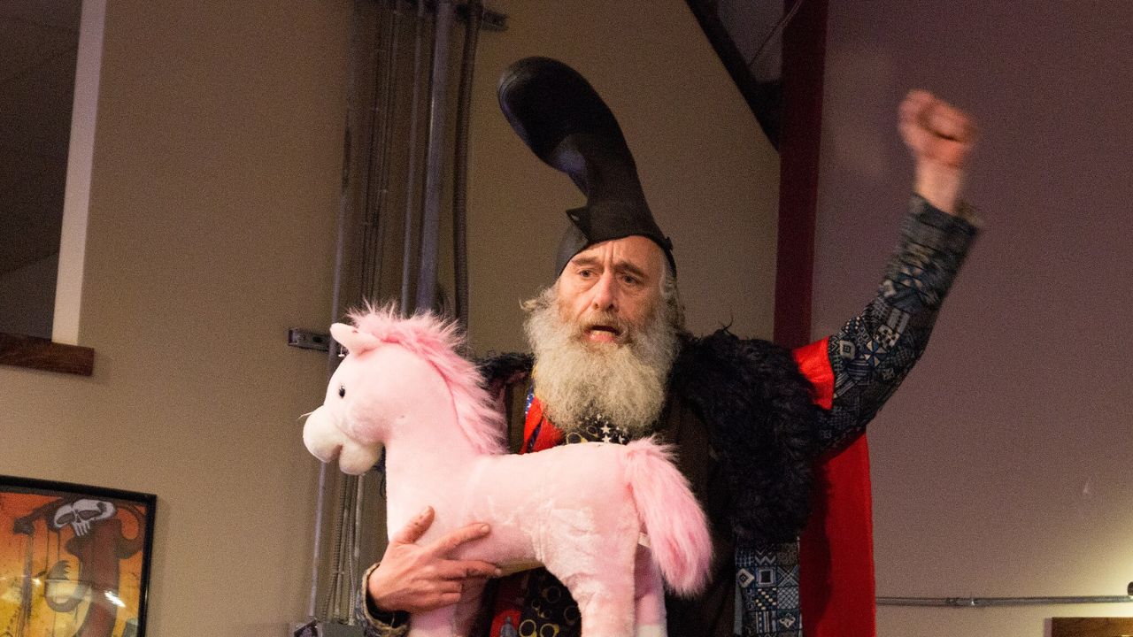 Vermin Supreme raising his hand in victory after a public appearance. He is wearing a large boot on his head and holding a stuffed pink unicorn with his other arm.