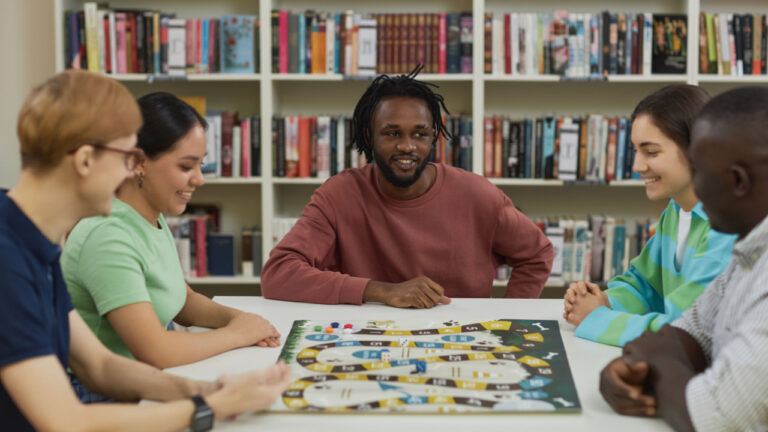 Diverse group of young people playing board games together while sitting around table in library and smiling