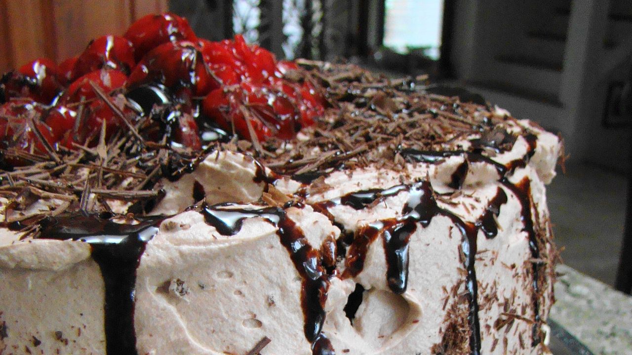 Black Forest Cake presented on a cake stand, fully topped with liquid chocolate and cherries.
