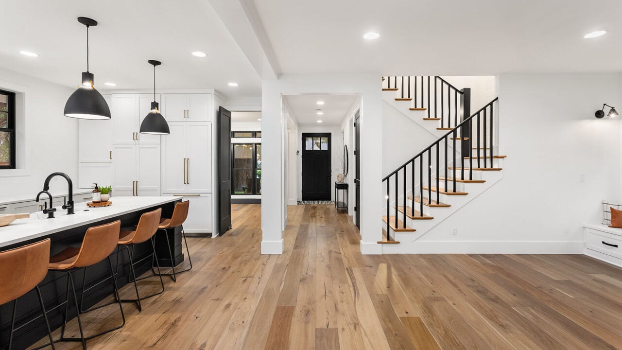 Kitchen and living room interior in new luxury home with open concept floor plan. Features large island with farmhouse sink, hardwood floors, and stairs to second story.