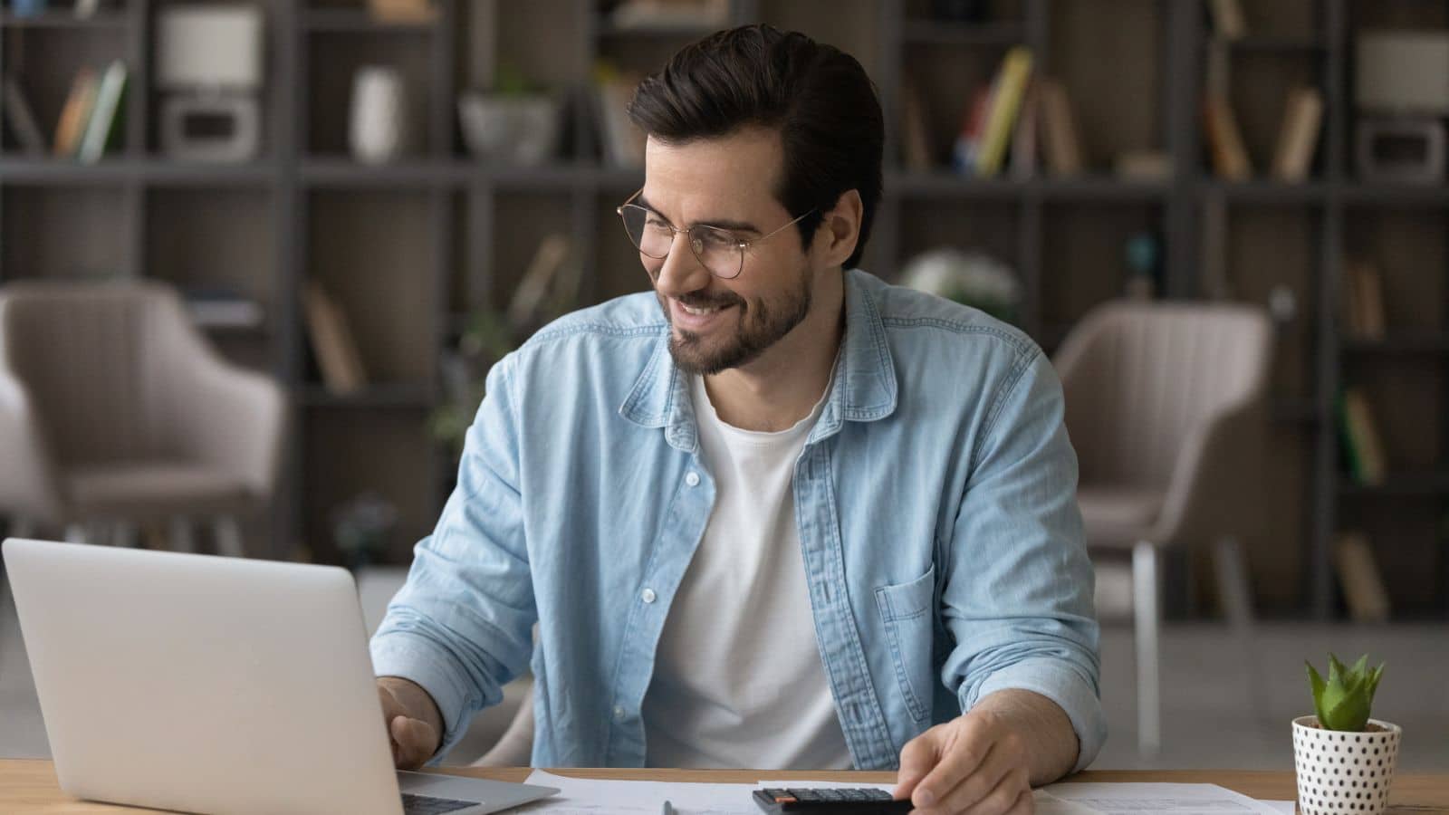 Smiling man in glasses using laptop, calculating bills, managing finances.