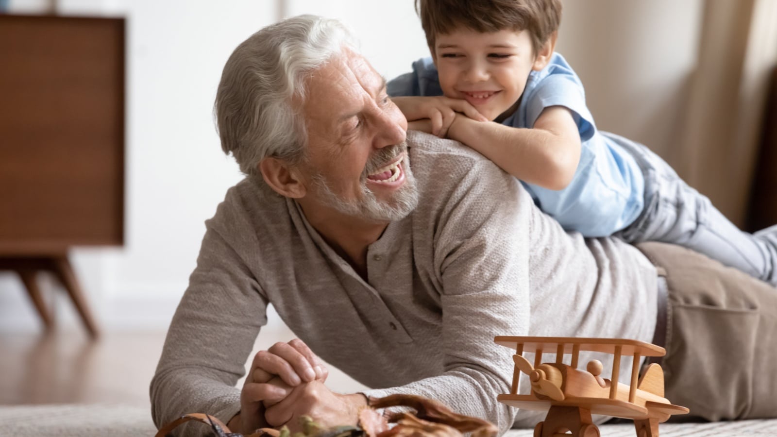Happy little boy lying on grandfather older man on floor carpet, having fun together in living room.