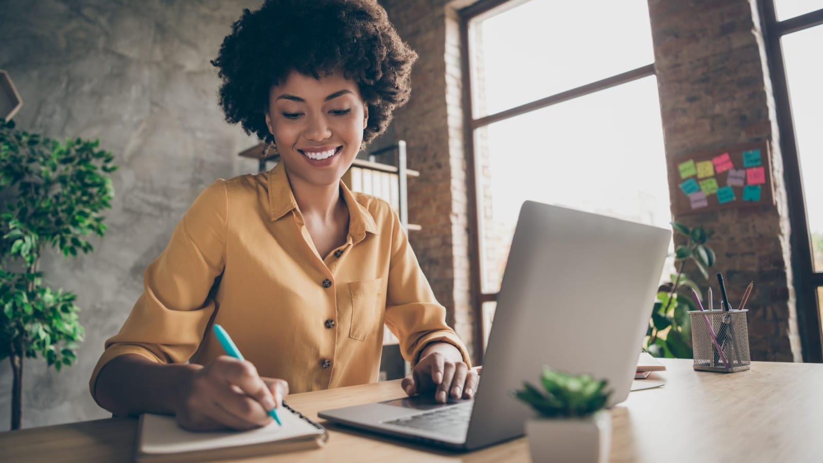 Happy woman smiling sitting at desktop with laptop writing down important information.