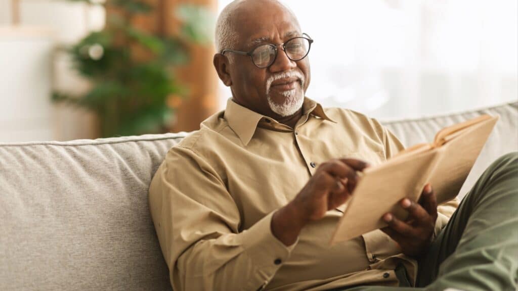 Image Credit: Senior African American Man Reading Book Sitting On Couch At Home.
