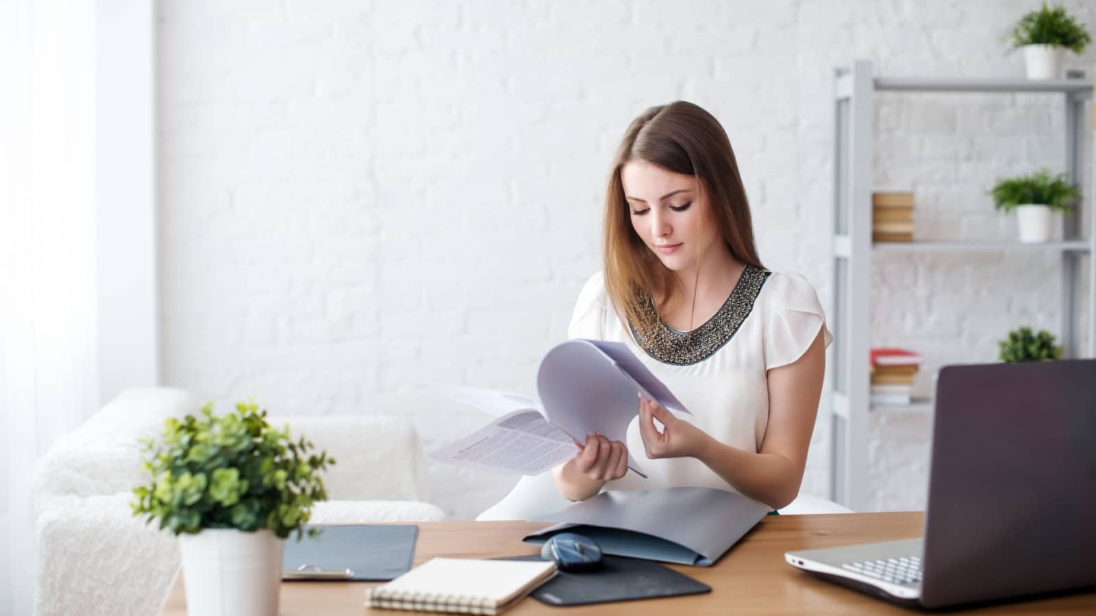 Woman flipping through papers at desk with plant on it.