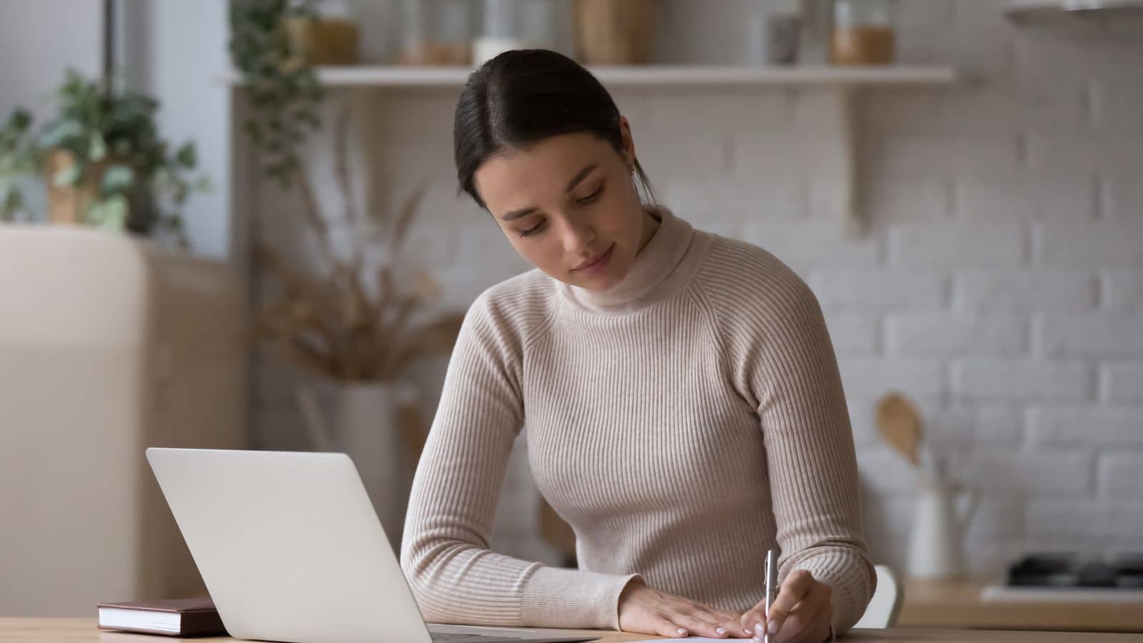 Woman sit at table with laptop jotting information on paper