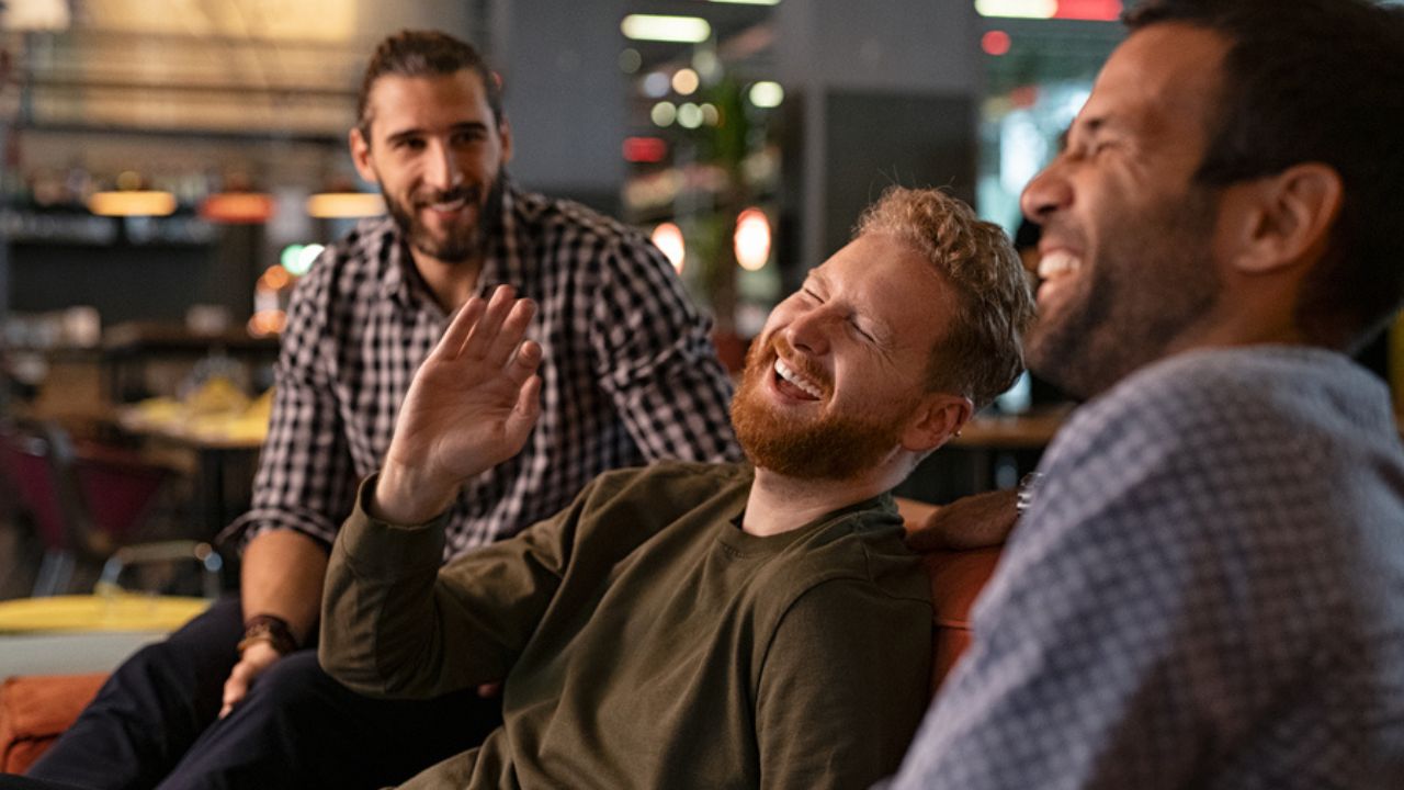 Group of three best friends laughing and enjoying the evening at pub. Happy young men enjoying late night staying together at bar. Cheerful guys sitting on couch and having fun while relaxing.