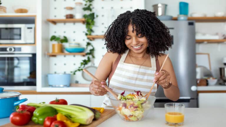 Close up portrait of happy cute beautiful young woman while she try tasty vegan salad in the kitchen at home.