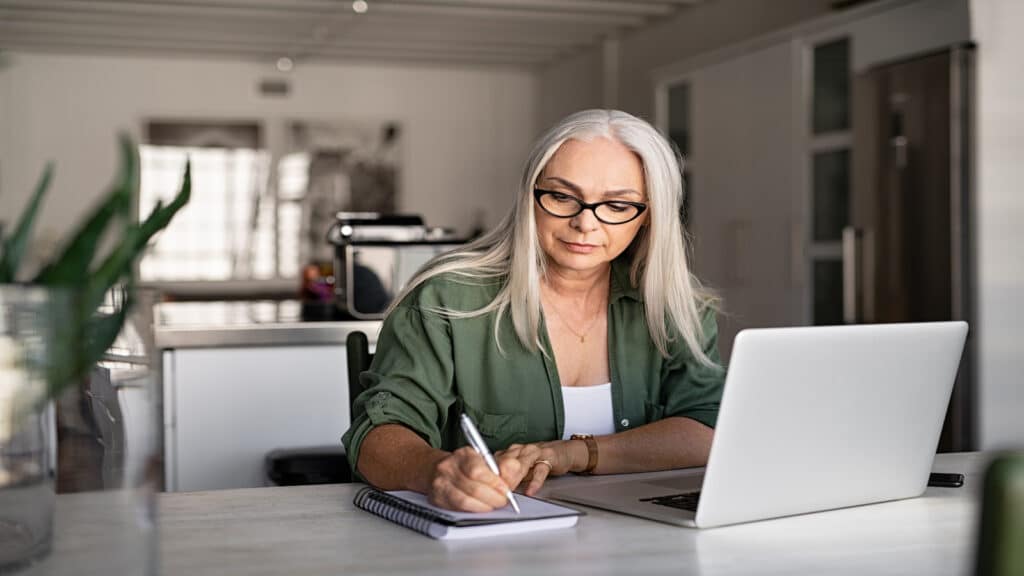 serious woman writing in notebook paying bills at computer.