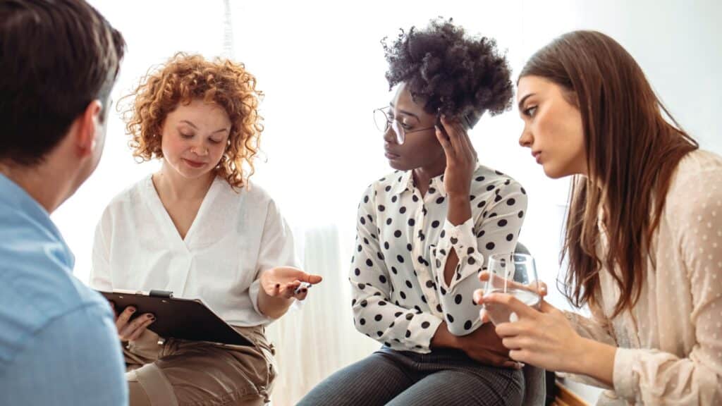 A diverse group of adults sit in a circle in their chairs and listen to and sympathize with each other with serious expressions. Men and women sitting in a circle during group therapy, talking.