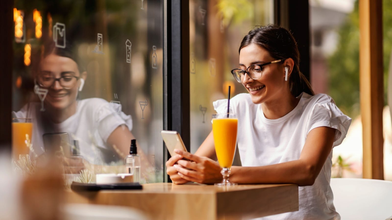 A young smiling woman is sitting alone in a restaurant on a coffee break, having a video call, and having a juice for drink.