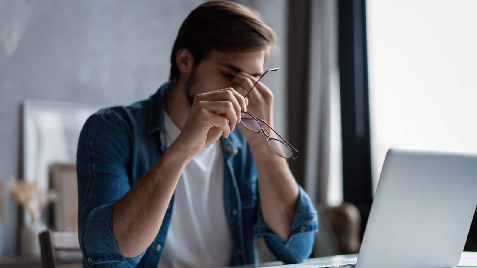 Frustrated young man massaging his nose and keeping eyes closed while sitting at his working place in office.