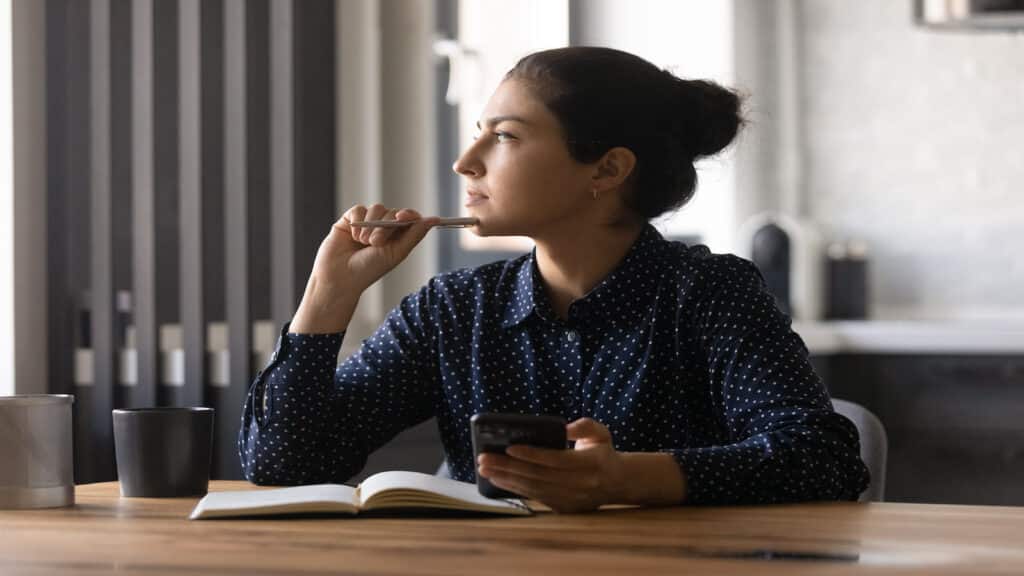 Pensive young female study by desk using mobile internet distracted from making notes create new idea.