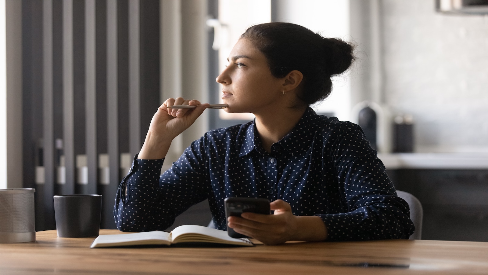 Pensive young female study by desk using mobile internet distracted from making notes create new idea.