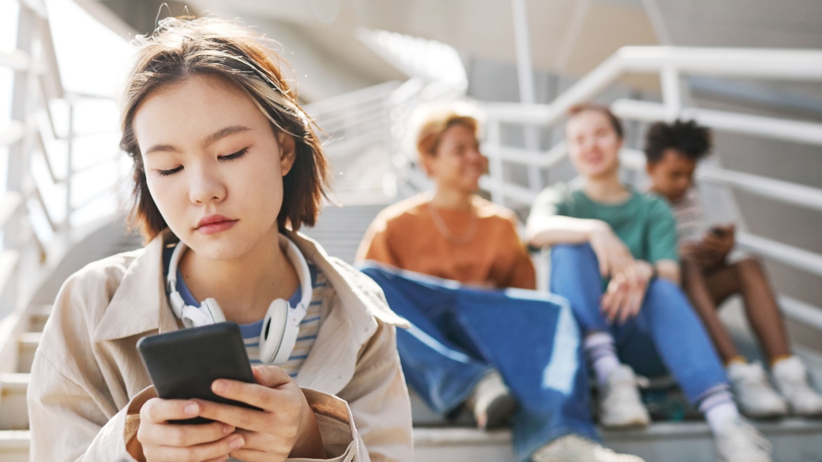 Portrait of Asian teenage girl holding smartphone outdoors while sitting on metal stairs with group of friends in background, copy space.
