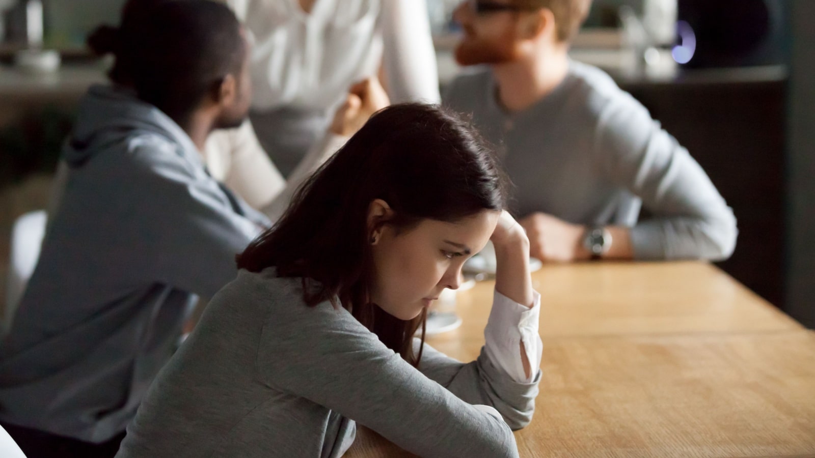 Sad frustrated young girl feeling lonely sitting alone at cafe table, upset social outcast or loner teenager suffering from low self esteem complex, unfair attitude or discrimination among friends.