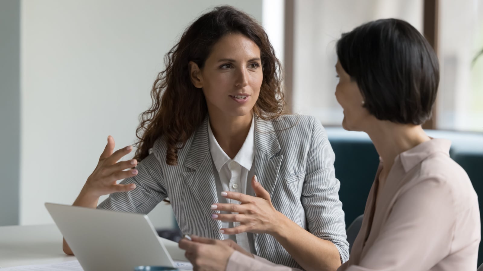 Two business women sit at desk discuss project details, diverse female colleagues met in office, share opinion, working on collaborative task, sales manager makes commercial offer to company client.