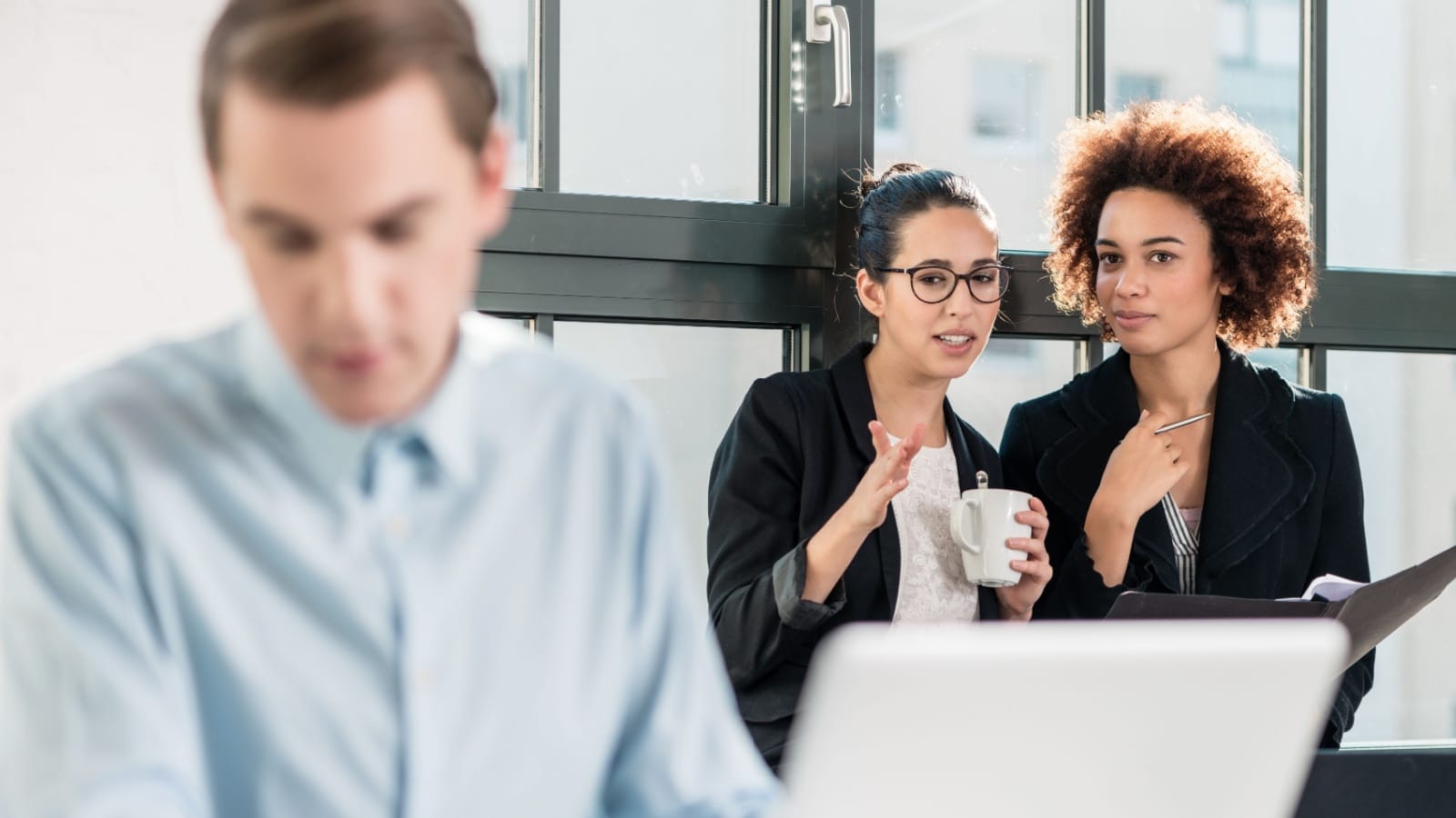 Two young cheerful female employees talking in the office next to their colleague working at desk.