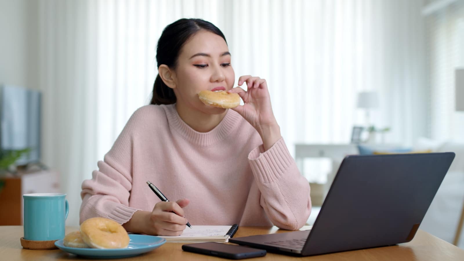 Young attractive beautiful asian female hungry eat doughnut take away snack food with full mouth look at computer notebook at home in busy work from home multitask unhealthy meal lifestyle concept.