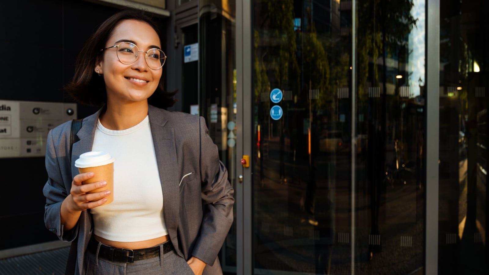 Young woman leaving the office building holding takeaway coffee and smiling.