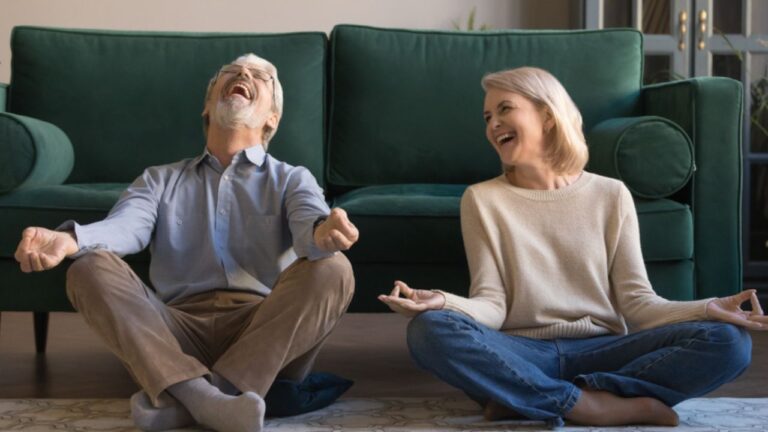 Laughing funny elderly spouses sitting in living room on floor in lotus position practice meditation distracted from yoga exercise joking feels overjoyed, healthy active lifestyle of retirees concept