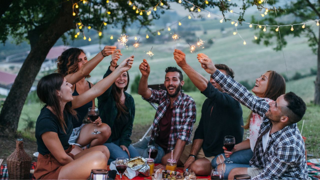 Group of happy millennials have fun with fire sparkles at picnic in spring at sunset on a camping holiday. People drinking wine and eating meat prepared at the barbecue