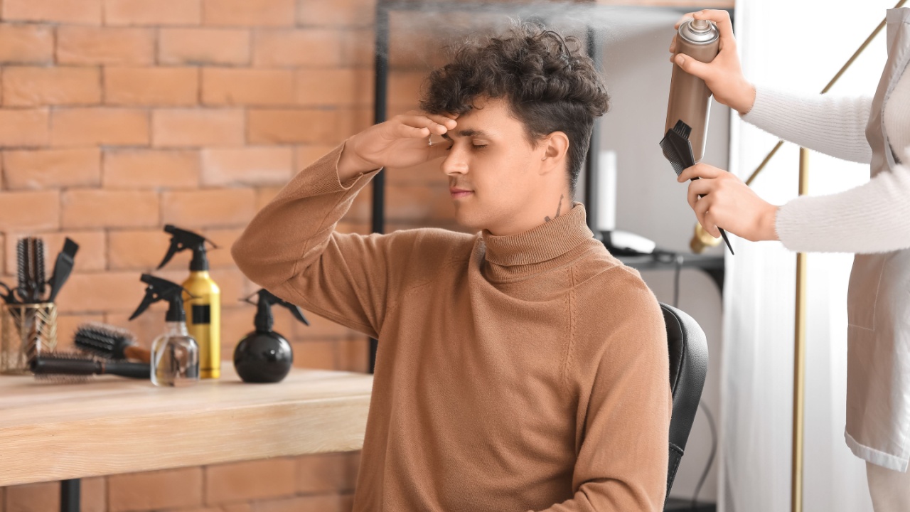 hairstylist applying hairspray as young man covers his face