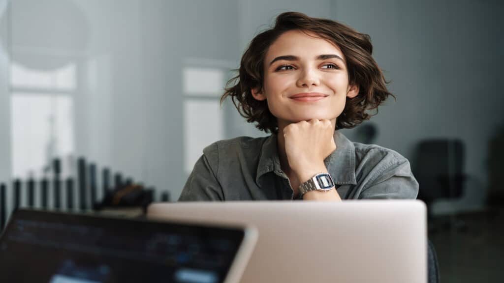 Image of young beautiful joyful woman smiling while working with laptop in office.