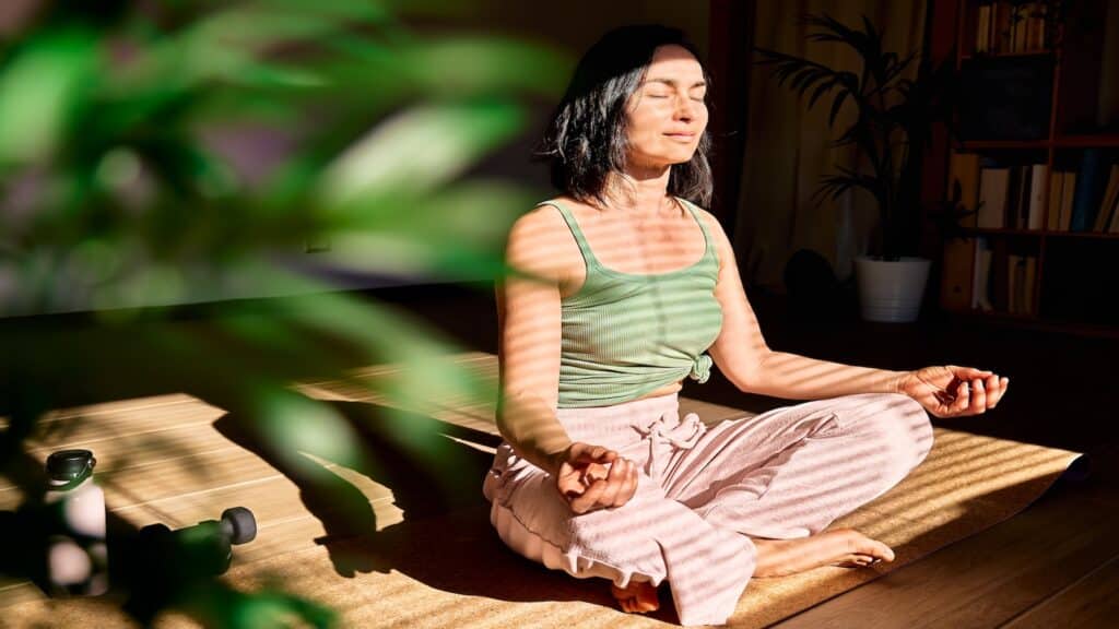 Woman practicing yoga and meditation at home sitting in lotus pose on yoga mat, relaxed with closed eyes.