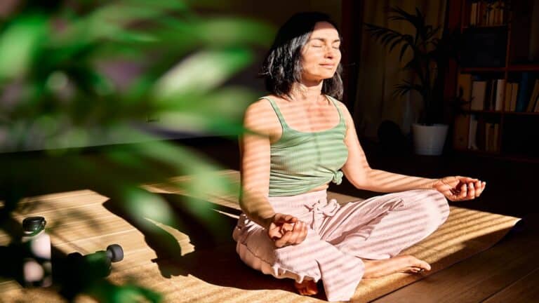 Woman practicing yoga and meditation at home sitting in lotus pose on yoga mat, relaxed with closed eyes.
