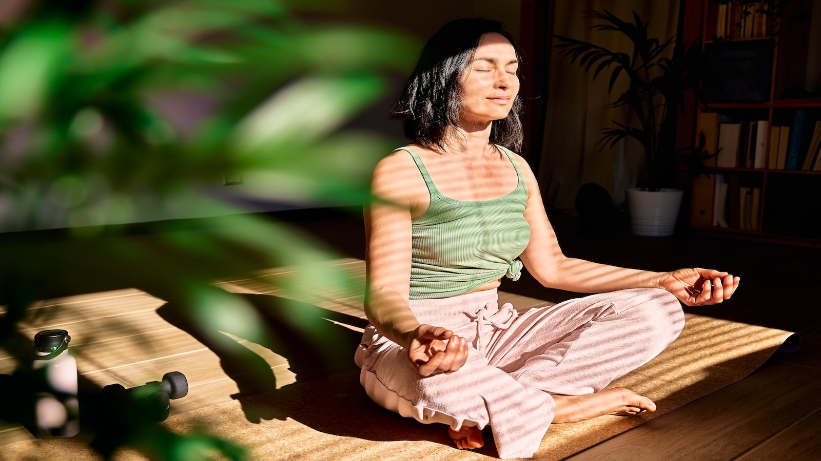 Woman practicing yoga and meditation at home sitting in lotus pose on yoga mat, relaxed with closed eyes.
