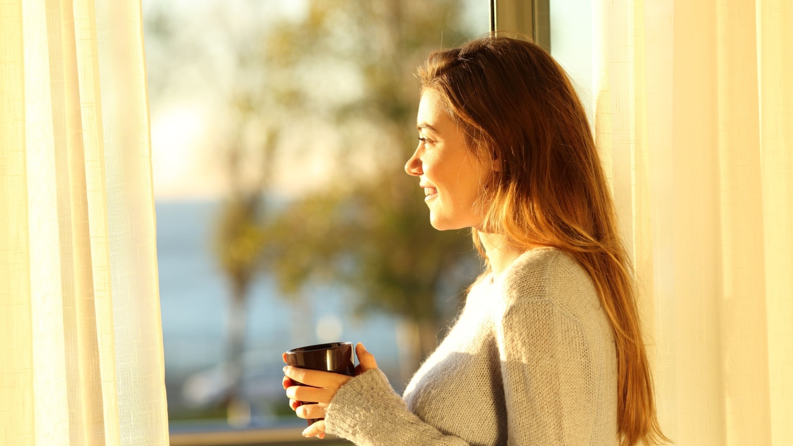 Woman drinking coffee watching sunrise from window smiling.