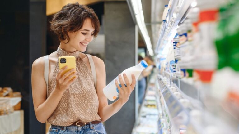 Young woman wear casual clothes backpack shopping at supermaket store buy choose dairy produce take milk read shelf life using mobile cell phone inside hypermarket Purchasing gastronomy food concept.