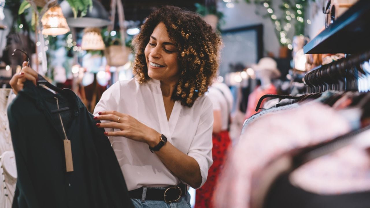 Smiling female standing near hangers with brand wear and laughing during Black Friday shopping with discounts, funny hipster girl enjoying pastime for update her wardrobe spending day in boutique