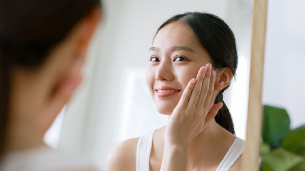 Young Asian woman touching healthy facial beauty skin look at mirror