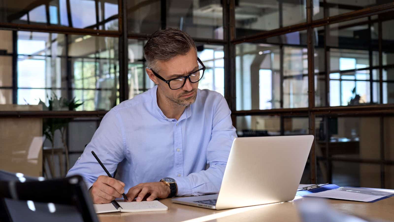 A man looking at a laptop and taking notes in an office.