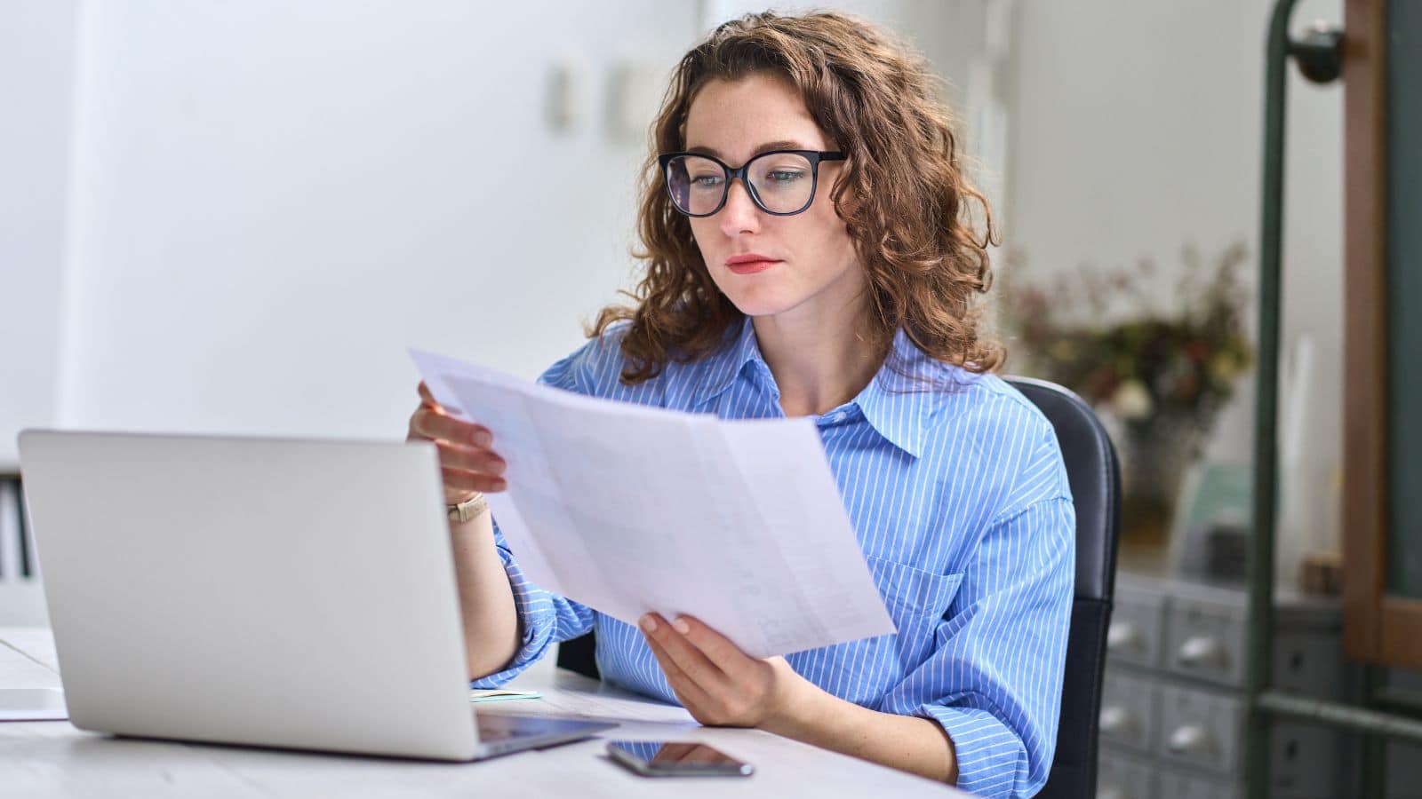 A woman holding a document working in an office with a laptop.