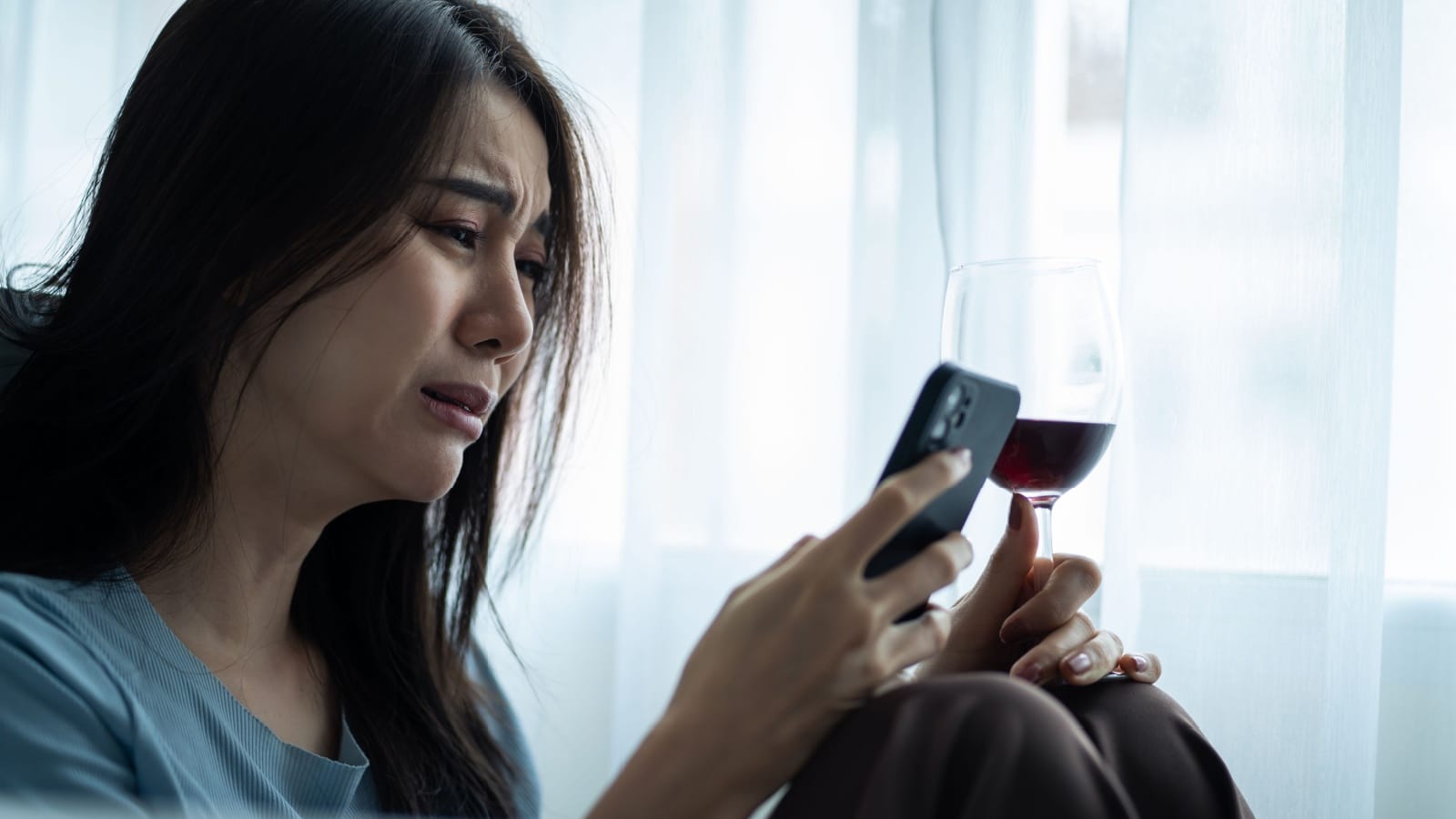 Asian young depression woman drinking wine while look at mobile phone. Attractive beautiful girl sitting on floor hold a bottle of alcohol feeling heart broken, drunk and hangover alone in living room.
