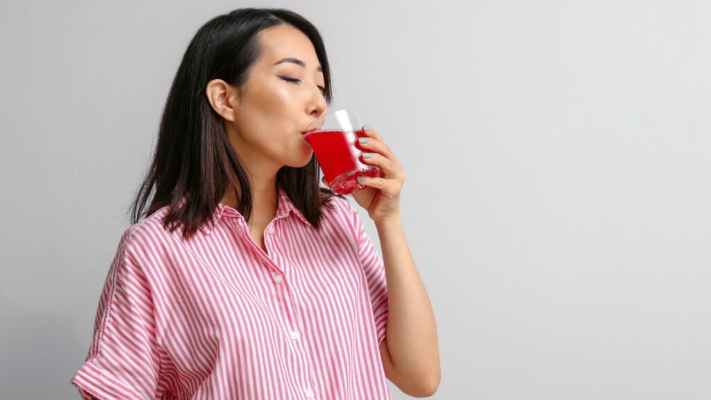Beautiful Asian woman drinking cherry juice on light background.