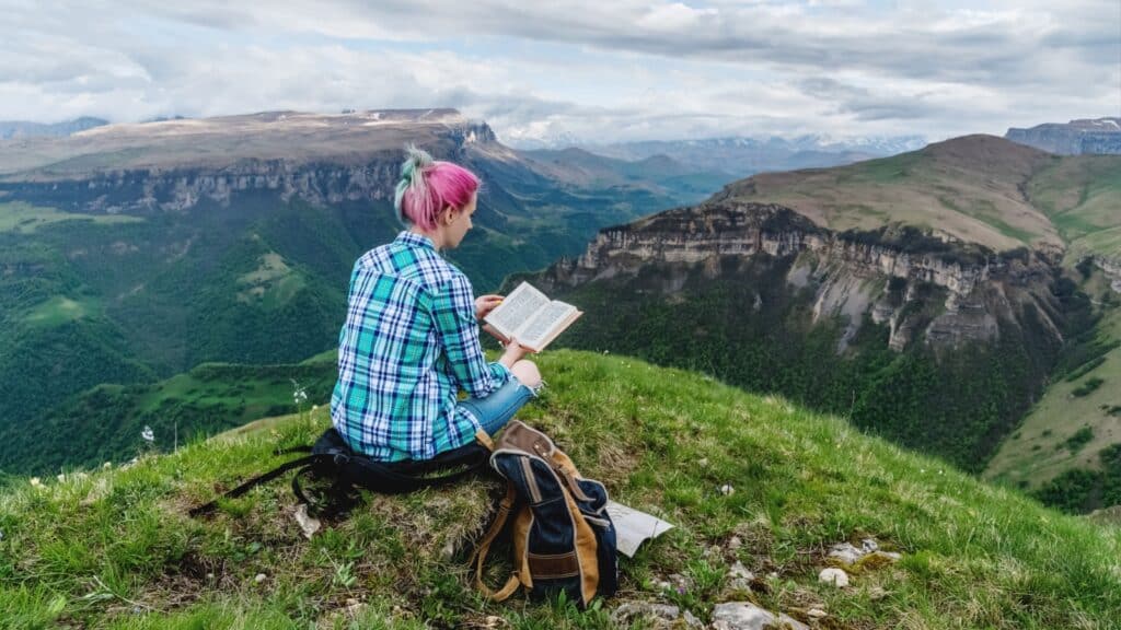 Beautiful young girl reads a book on a background of beautiful mountains. The concept of hanging out, relaxing and working
